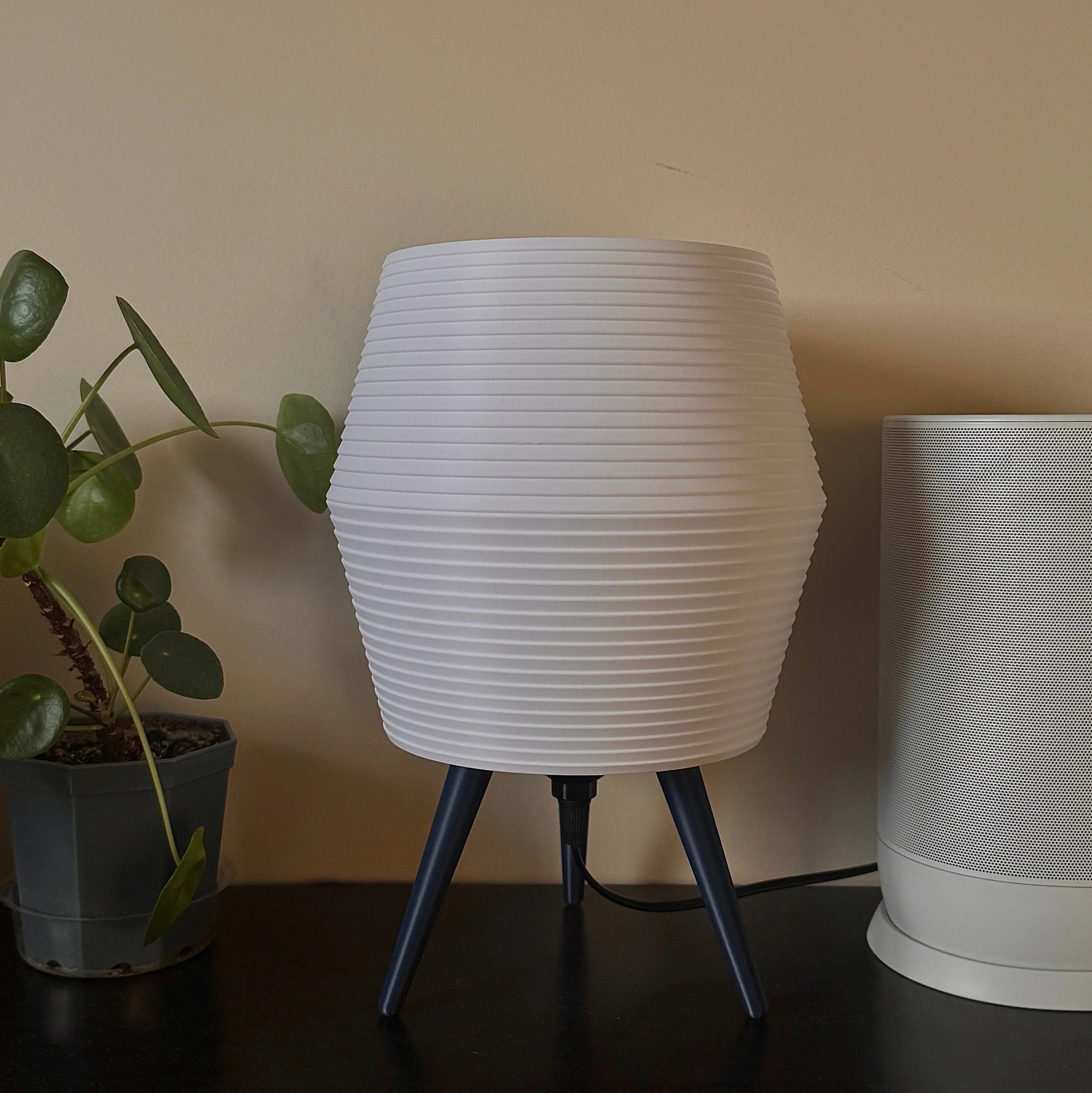 White textured table lamp with blue legs on a dark surface next to a plant.