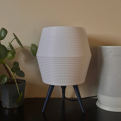 White textured table lamp with blue legs on a dark surface next to a plant.