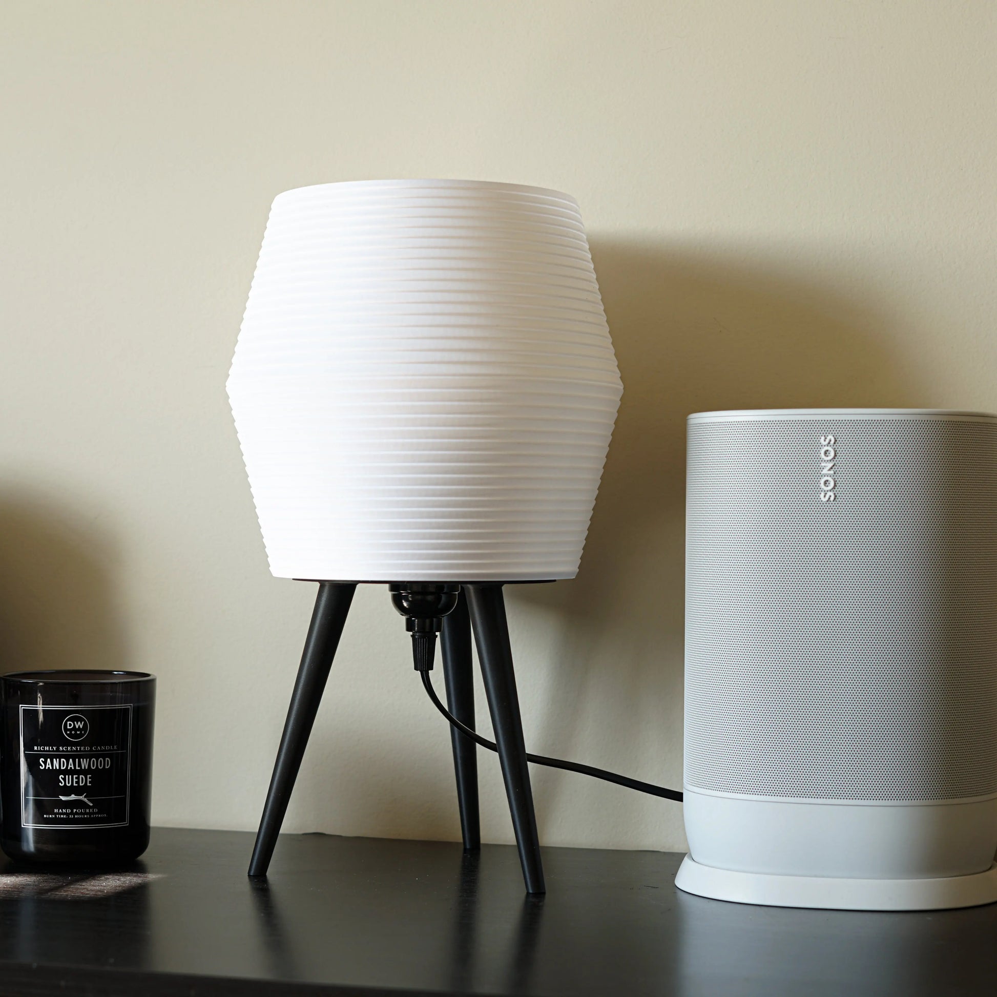 White lamp with black legs and a white Sonos speaker on a dark surface against a beige wall.