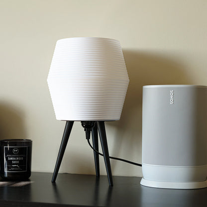 White lamp with black legs and a white Sonos speaker on a dark surface against a beige wall.