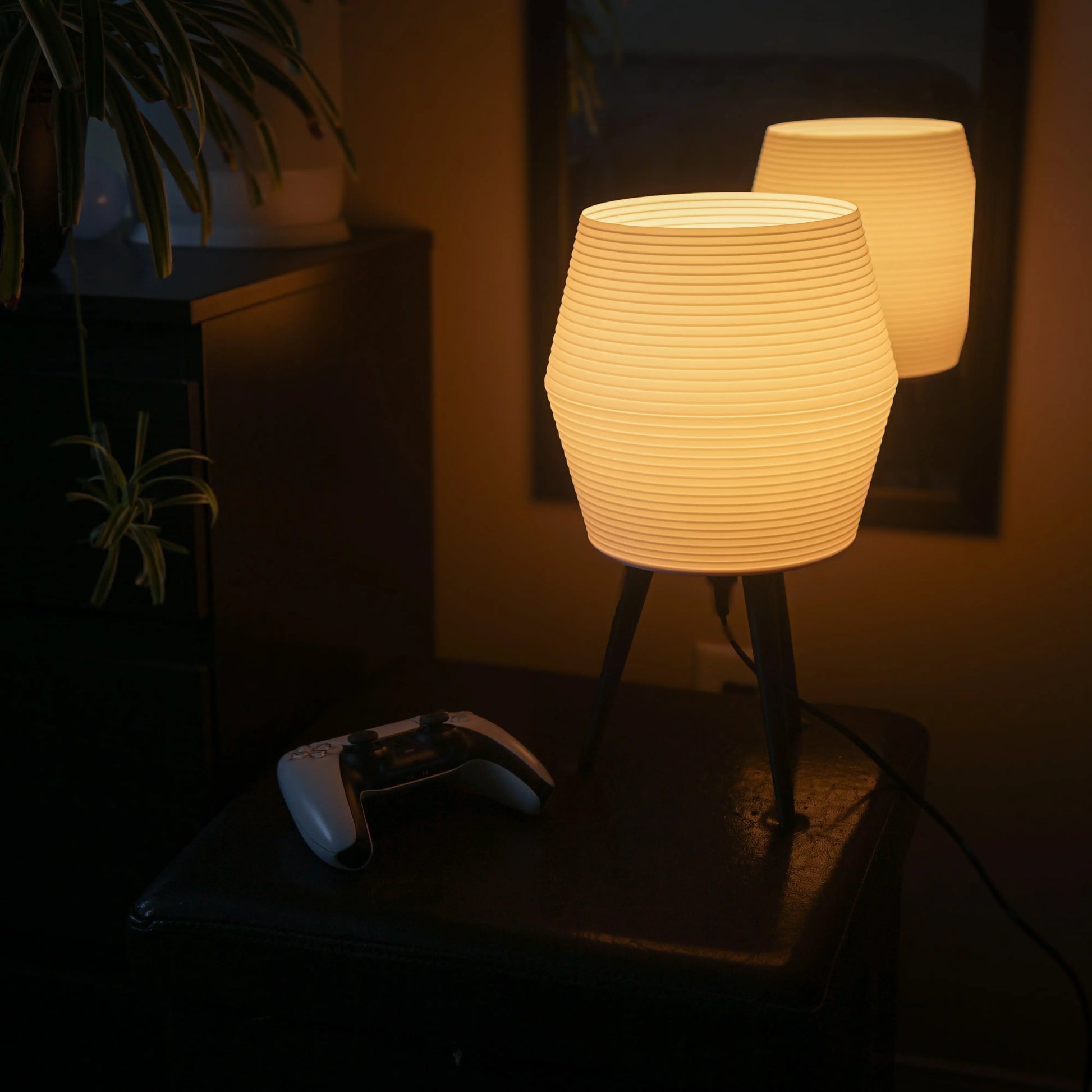 Lamp emitting a warm glow on a night stand with a playstation controller next to it.